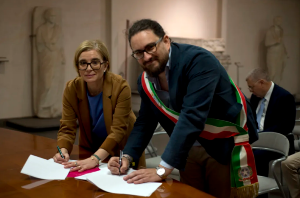 Two officials sign documents at a formal ceremony, with a man wearing an Italian-tricolor sash leaning over the table next to a woman in a brown blazer helping with paperwork in a stately room.