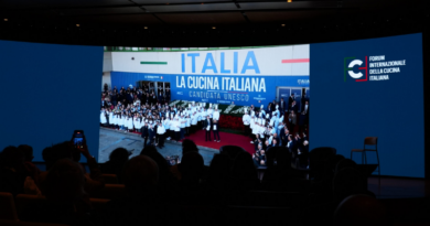 Stage at an Italian culinary forum showing a crowd of chefs in white jackets on red carpet beneath a banner reading 'ITALIA La CUCINA ITALIANA' with audience silhouettes in the foreground.