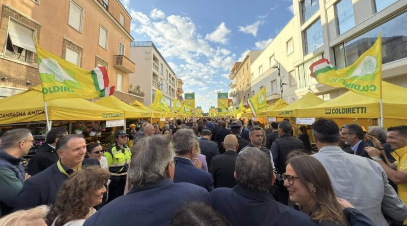 Crowded street market with bright yellow Coldiretti tents and flags lining both sides under a blue sky. People mingle among vendors and stalls.